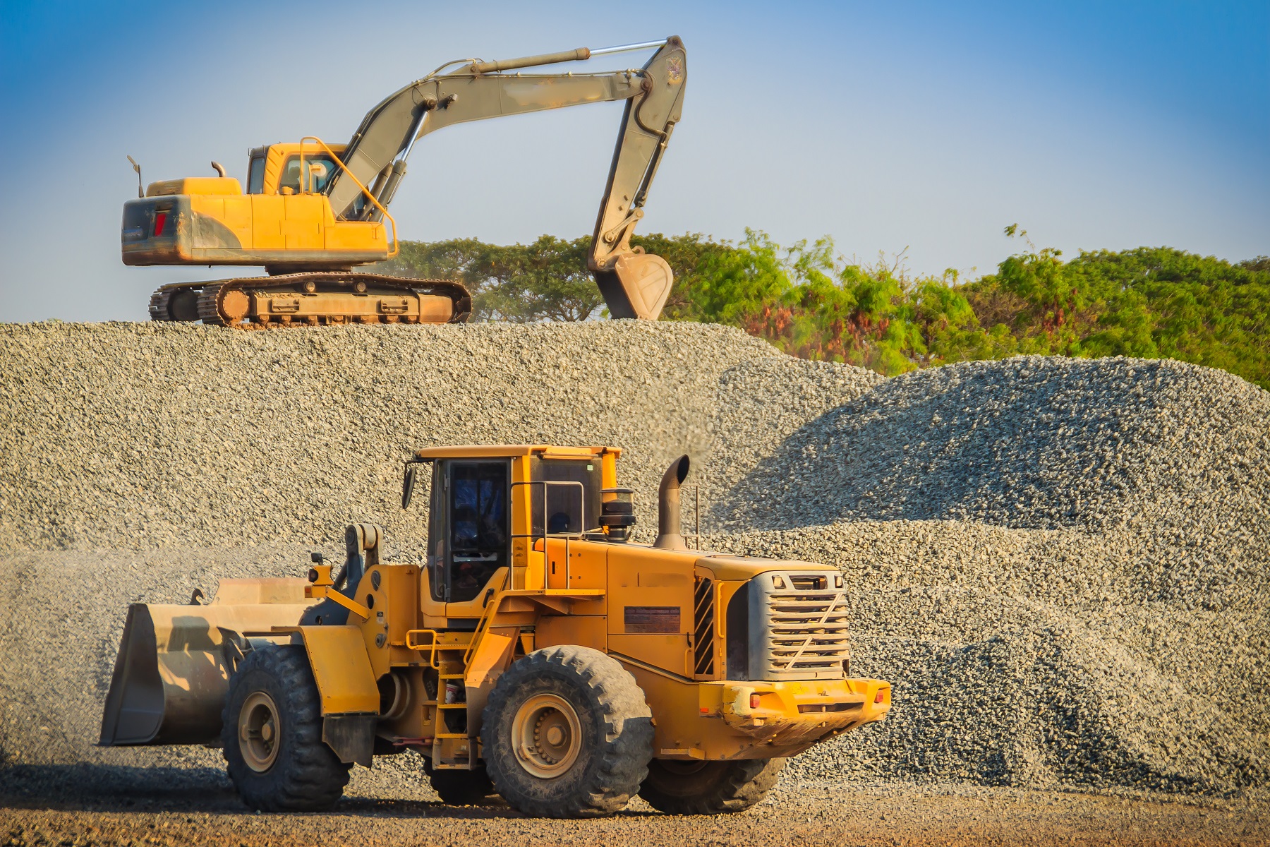 Yellow wheel loader and excavator are working in quarry against the background of crushed stone storage.